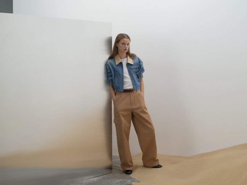 A woman poses in a light blue denim jacket, white t-shirt, and tan wide-leg pants, set against a minimal background with a neutral gradient.