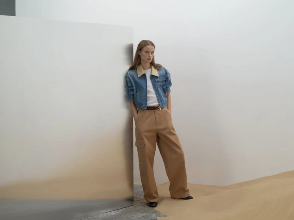 Model leaning against a metal wall in a denim jacket and tan trousers for Sandro S/S 25 Lookbook