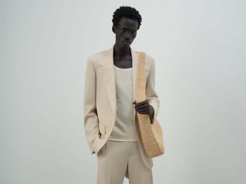 Male model in a beige suit and tank top, carrying a tan raffia hobo bag against a plain background in a fashion photo studio.