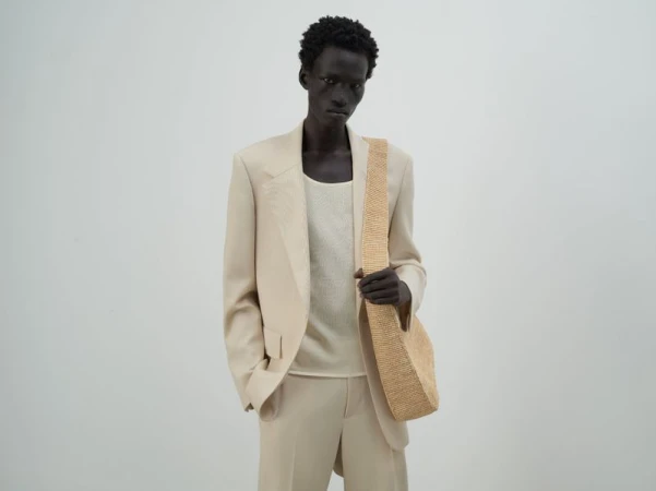 Male model in a beige suit and tank top, carrying a tan raffia hobo bag against a plain background in a fashion photo studio.