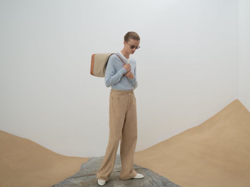 Adult female model wears light blue sweater and beige pants with a shoulder bag posing in a bright studio with minimal set design.