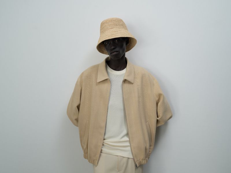 Dark-skinned male model in a tan jacket, t-shirt, pants, and a straw bucket hat posing in front of a plain gray backdrop.