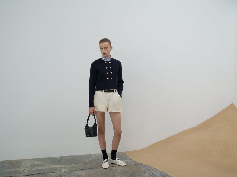 A woman poses in a navy jacket, white shorts, and black socks with white shoes in a minimalist setting with a modern aesthetic.