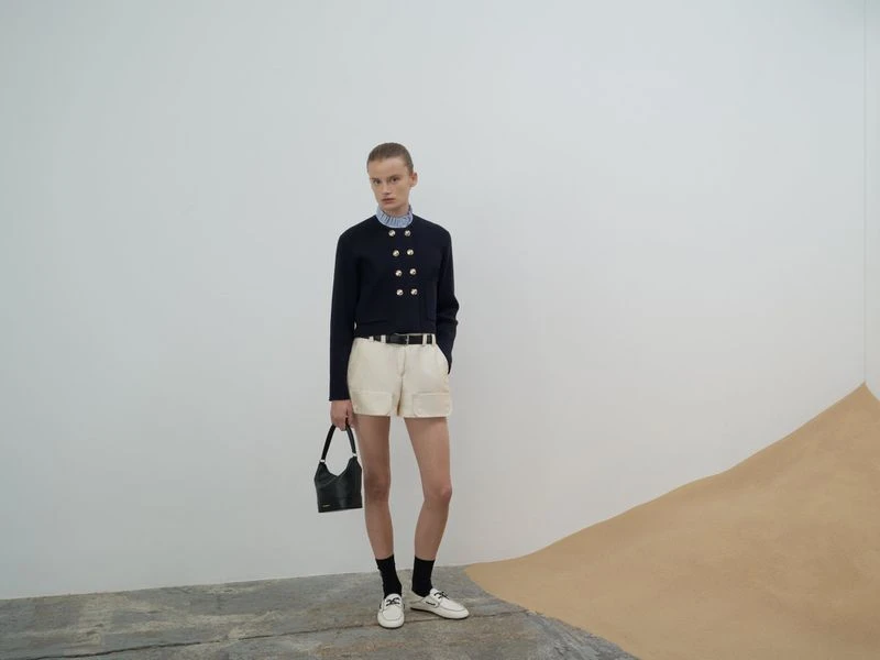 A woman poses in a navy jacket, white shorts, and black socks with white shoes in a minimalist setting with a modern aesthetic.