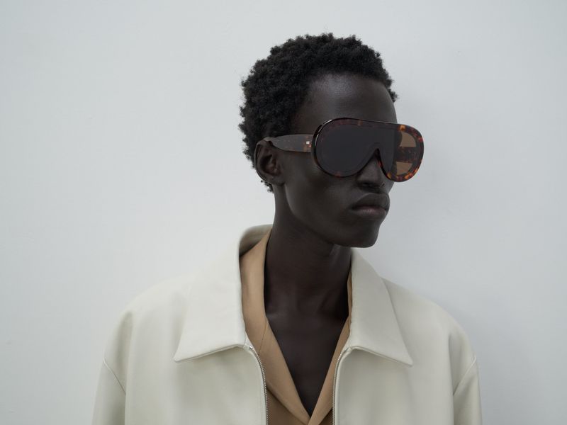 Studio shot of a person wearing large sunglasses, a beige jacket, and a light tan shirt in front of a plain, light grey background.