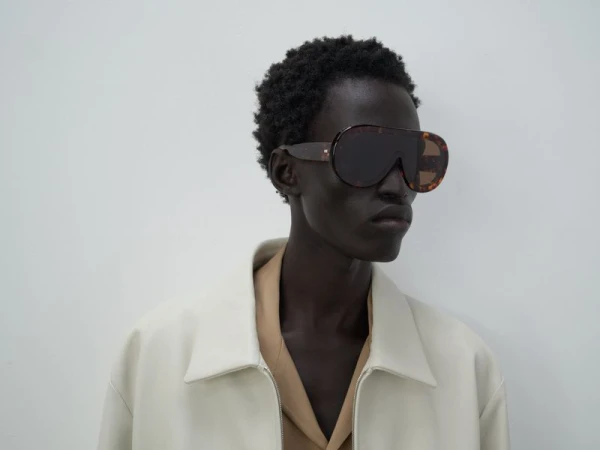 Studio shot of a person wearing large sunglasses, a beige jacket, and a light tan shirt in front of a plain, light grey background.