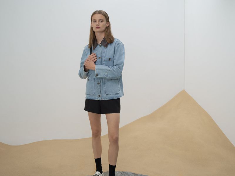 Woman in denim jacket and black shorts poses in a studio environment with a sand dune backdrop against a white wall.