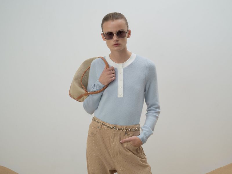 A woman in sunglasses, a light blue sweater, and tan pants poses with a tan purse against a neutral background in a fashion studio shot.