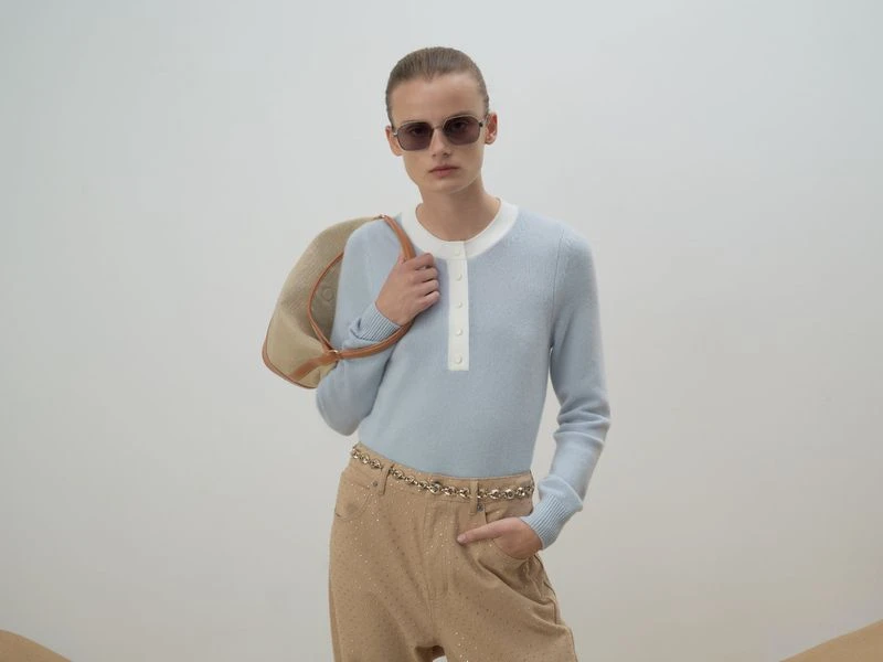 A woman in sunglasses, a light blue sweater, and tan pants poses with a tan purse against a neutral background in a fashion studio shot.