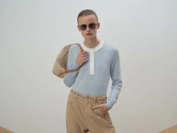 A woman in sunglasses, a light blue sweater, and tan pants poses with a tan purse against a neutral background in a fashion studio shot.