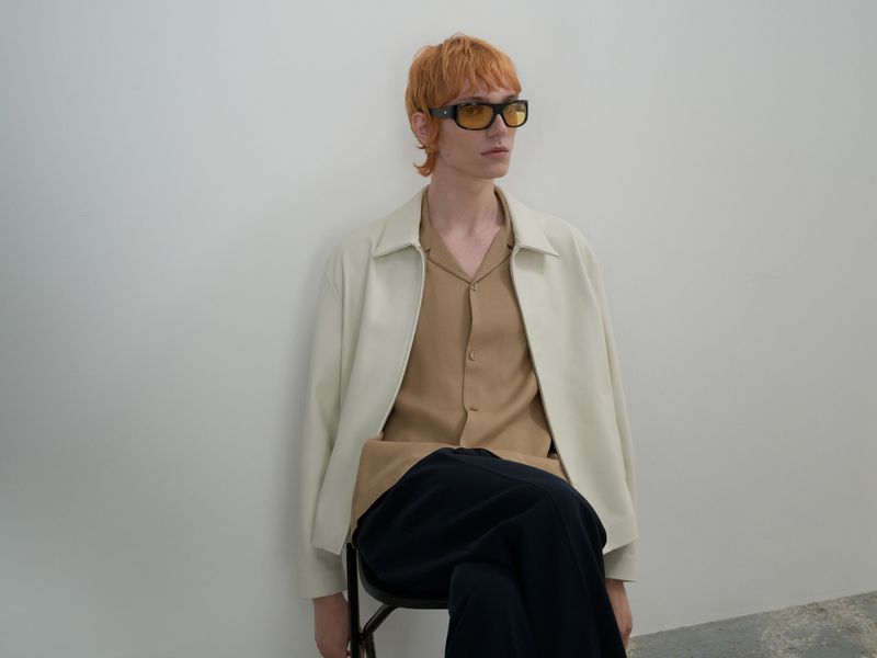 Adult male with red hair and yellow sunglasses wearing a tan collared shirt and cream jacket sitting in a white indoor studio.