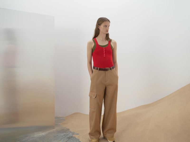 A slender woman poses in a red tank top and khaki cargo pants against a backdrop of a minimalist set featuring tan-colored sand.