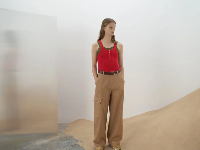 A slender woman poses in a red tank top and khaki cargo pants against a backdrop of a minimalist set featuring tan-colored sand.