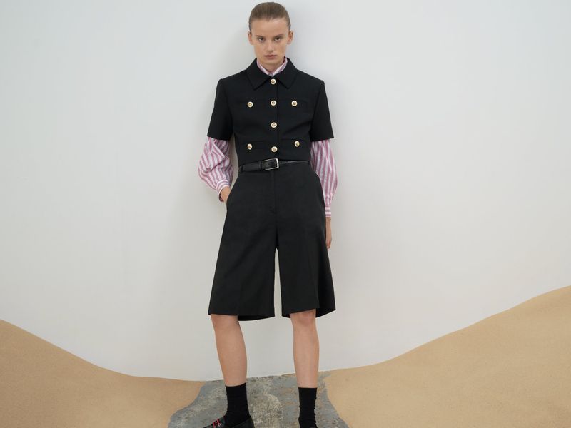 Fashionable woman in black short suit with gold buttons, pink striped top, black socks and platform shoes posing against white backdrop.