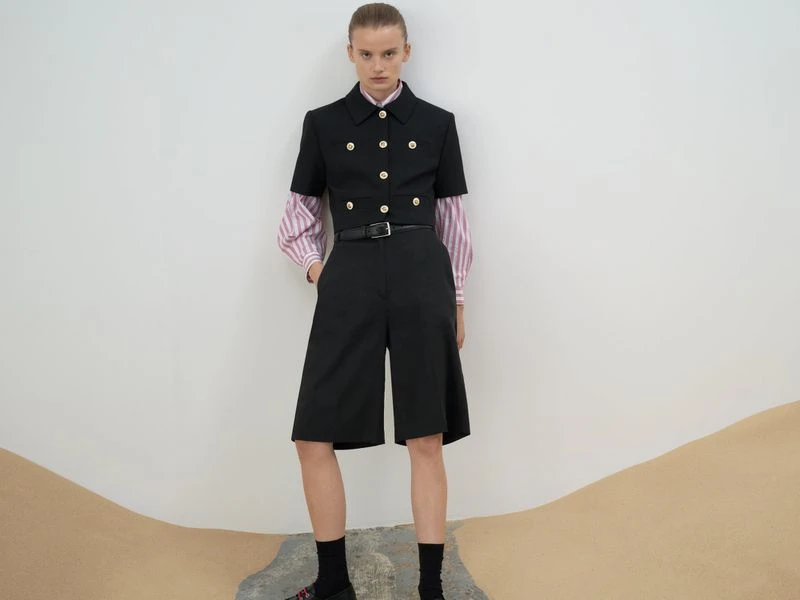 Fashionable woman in black short suit with gold buttons, pink striped top, black socks and platform shoes posing against white backdrop.