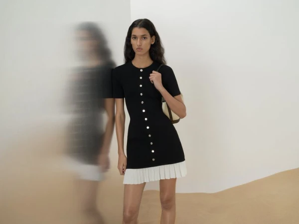 Model wearing a black buttoned mini dress with white pleated hem against a neutral background with motion blur figure