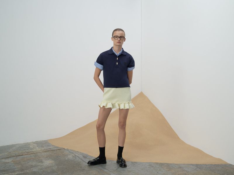 Adult fashion model poses in a studio wearing a navy polo shirt with a light yellow skirt, black socks, and black loafers.