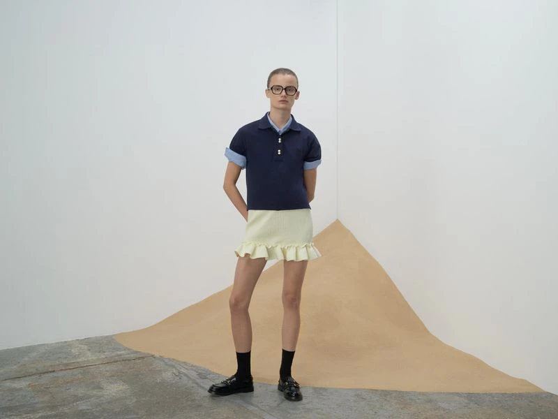Adult fashion model poses in a studio wearing a navy polo shirt with a light yellow skirt, black socks, and black loafers.