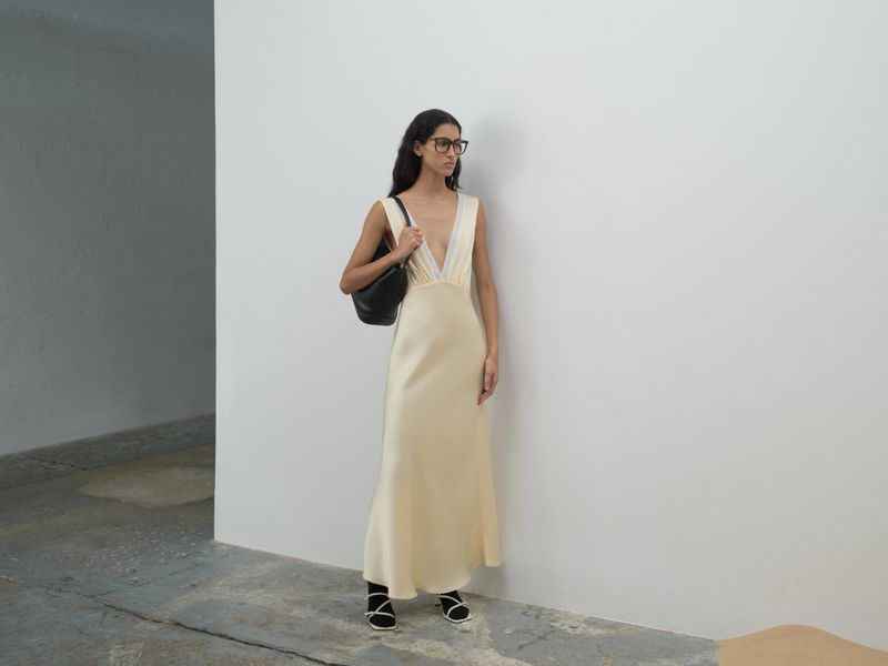 Model wearing a pale yellow slip dress and black handbag standing against a white wall.