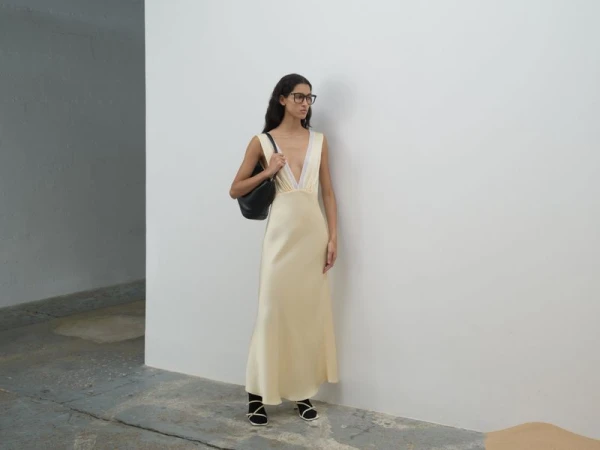 Model wearing a pale yellow slip dress and black handbag standing against a white wall.