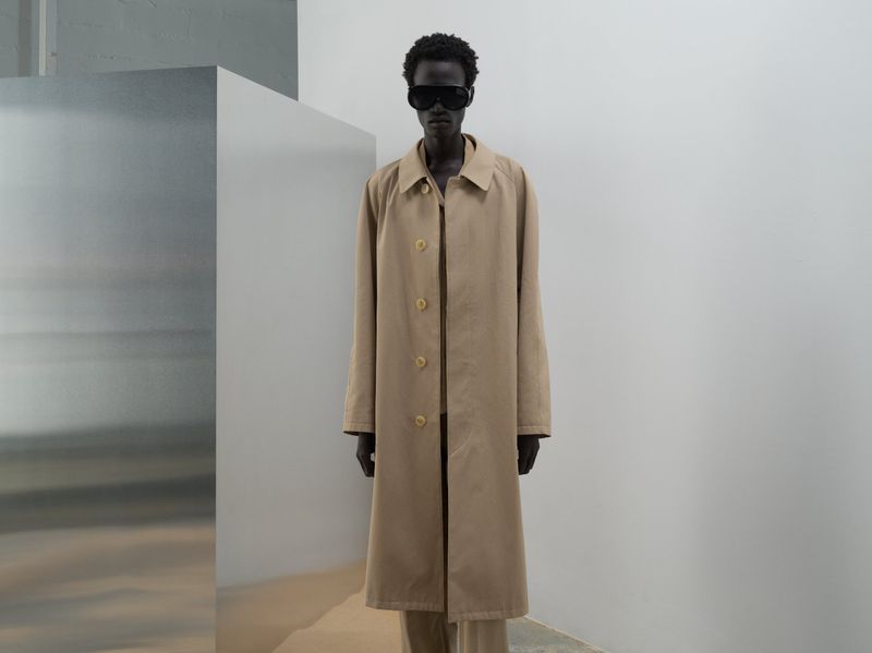 Fashionable adult in a beige coat and sunglasses stands in front of a minimalist background, showcasing a blend of neutral tones and textures.