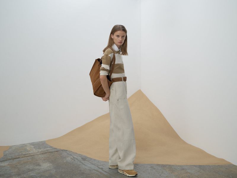 Fashion shot of a woman in wide-legged pants and a striped shirt posing indoors with a backpack against a neutral backdrop.