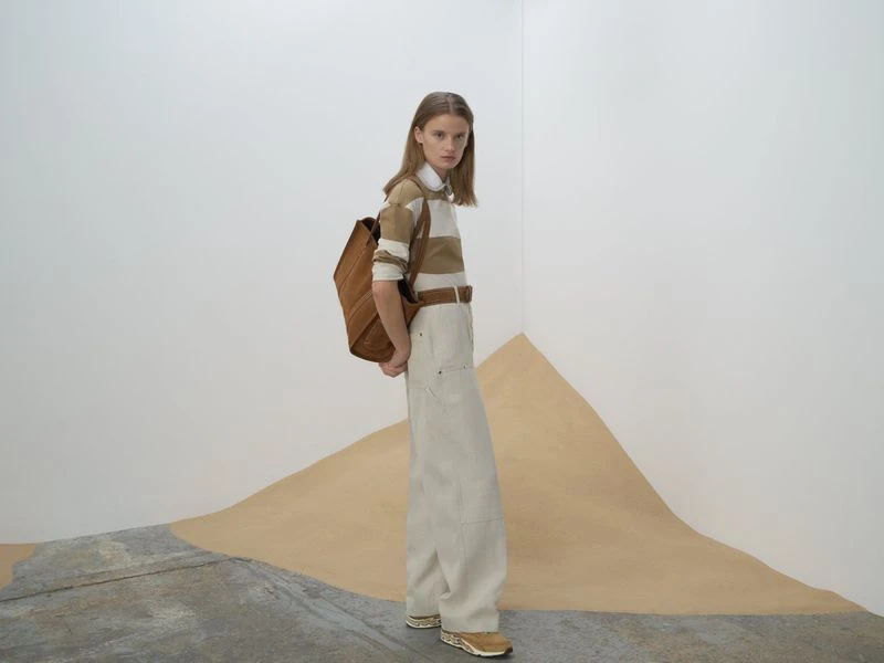 Fashion shot of a woman in wide-legged pants and a striped shirt posing indoors with a backpack against a neutral backdrop.