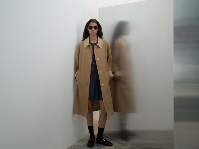 A young woman poses in a tan trench coat and pinstripe dress next to a frosted glass pane in a minimalist studio with neutral light.