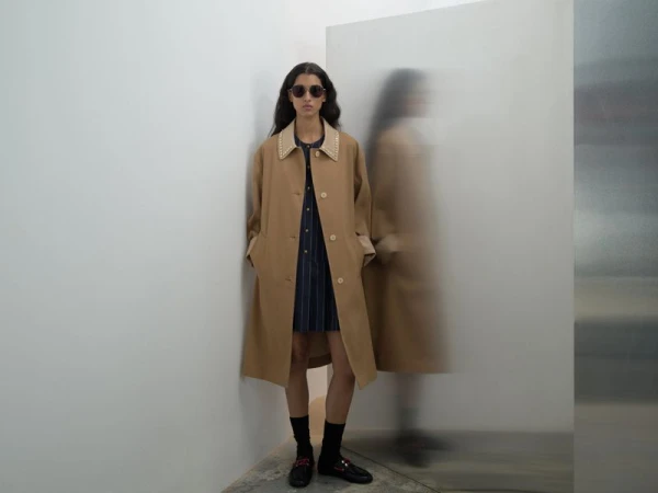 Model in dark sunglasses wearing a tan coat and dark dress against a white studio wall with a motion-blurred reflection