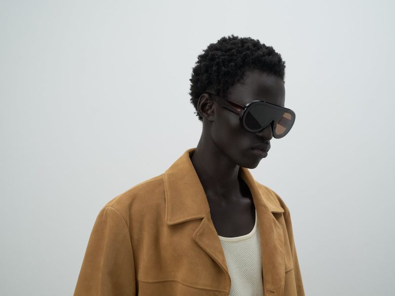 Dark-skinned adult male model poses indoors wearing sunglasses and a brown jacket against a simple grey background, fashion style.