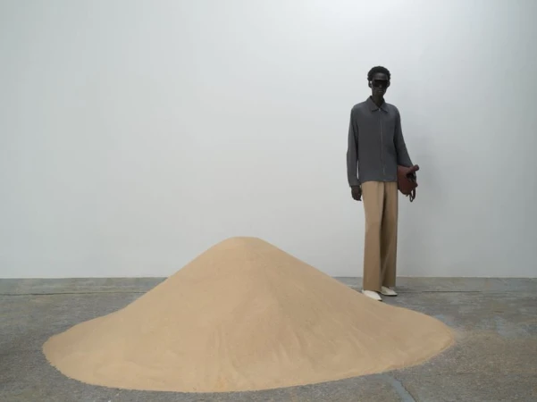 Model standing behind a large mound of sand in a studio wearing a grey jacket, tan trousers, and sunglasses