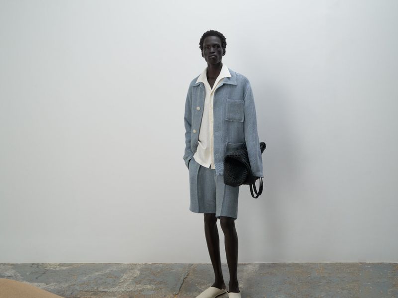 A dark-skinned man in a light blue linen suit with an off-white shirt poses in front of a white wall, holding a black woven bag.