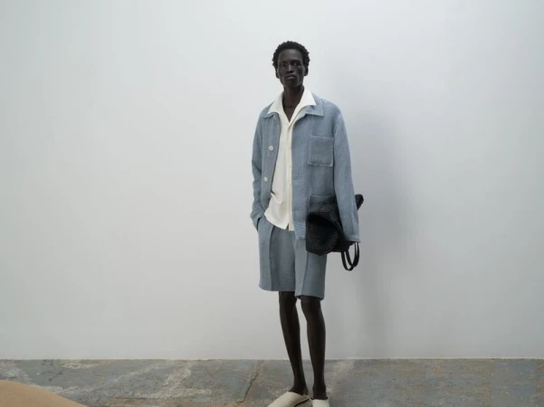 A dark-skinned man in a light blue linen suit with an off-white shirt poses in front of a white wall, holding a black woven bag.