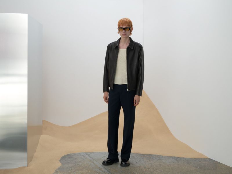 Male fashion model poses in an indoor photo studio wearing a dark leather jacket, white tank top, dark pants, and yellow tinted sunglasses.