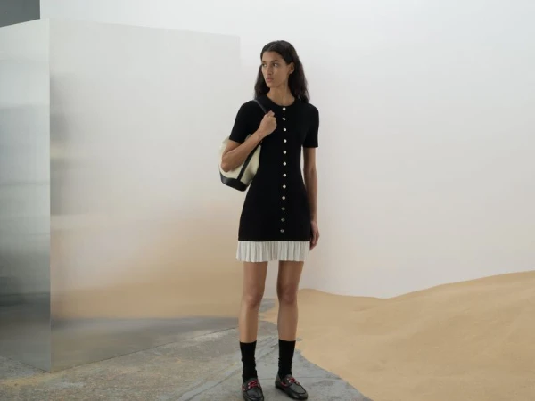 Model wearing a black button-front dress and carrying a shoulder bag against a white wall with sand on the floor.