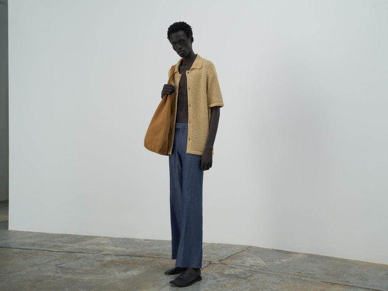 An adult male model poses in a photo studio wearing a knitted tan shirt, loose blue pants, and a tan tote bag.