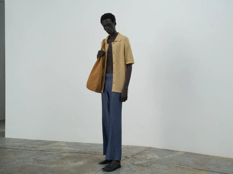 An adult male model poses in a photo studio wearing a knitted tan shirt, loose blue pants, and a tan tote bag.