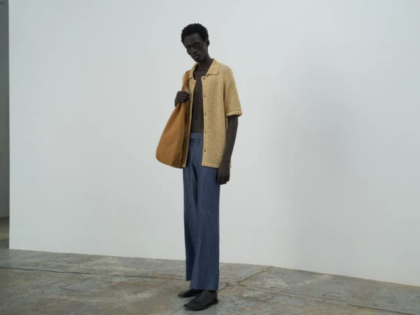 An adult male model poses in a photo studio wearing a knitted tan shirt, loose blue pants, and a tan tote bag.