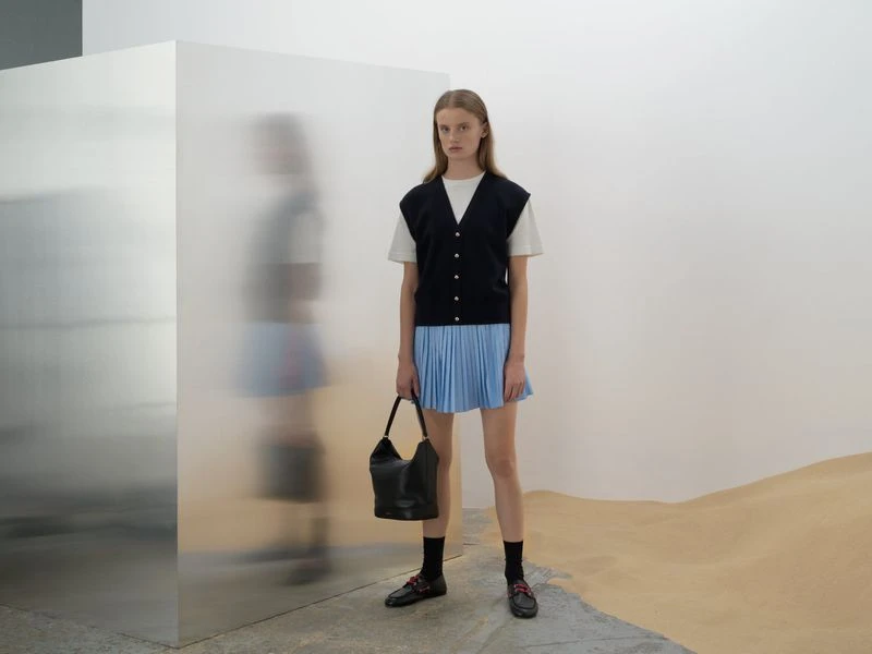 A young woman in a navy vest and light blue pleated skirt stands in a minimalist studio holding a black leather bucket bag.