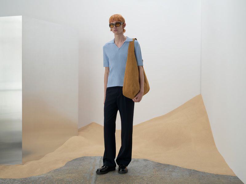 A stylish redhead adult in a blue ribbed polo shirt stands indoors with a brown tote bag and black trousers against a backdrop of painted dunes.