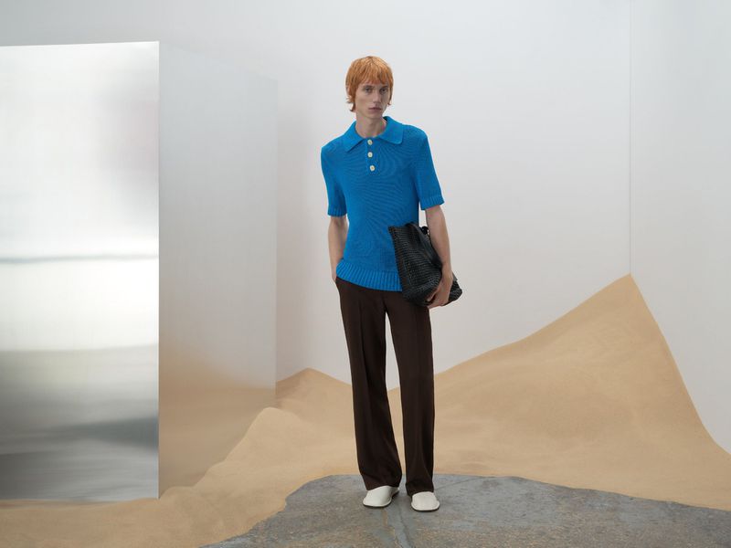 A slender man with red hair poses indoors wearing a blue short-sleeved collared shirt, brown slacks, and white shoes while holding a dark tote.