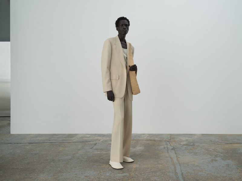 A fashion shot of a dark-skinned model wearing a beige suit with white shoes, standing in a bright studio with a woven bag.