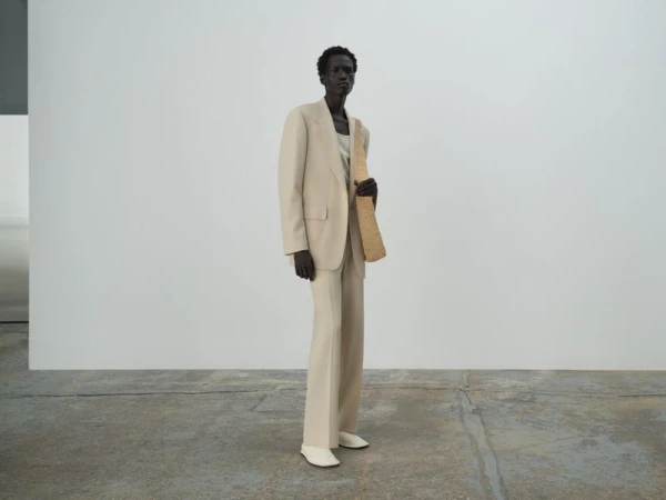 A fashion shot of a dark-skinned model wearing a beige suit with white shoes, standing in a bright studio with a woven bag.