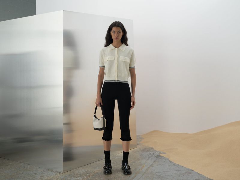 Indoor shot of a young adult female fashion model in a white top and black pants, posing with a white handbag near a metal cube.