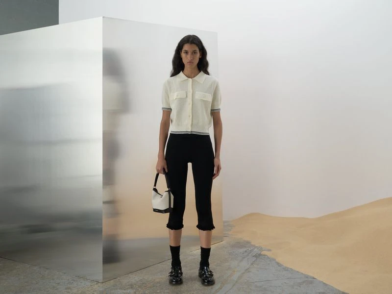 Indoor shot of a young adult female fashion model in a white top and black pants, posing with a white handbag near a metal cube.