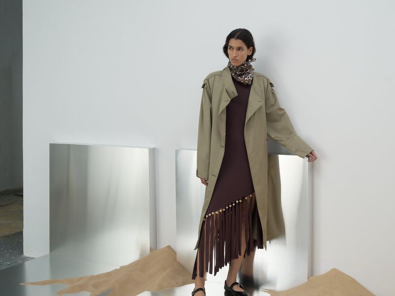 Fashion shot of a woman in a brown dress and tan trench coat standing in a studio with reflective decor and sand details on the floor.