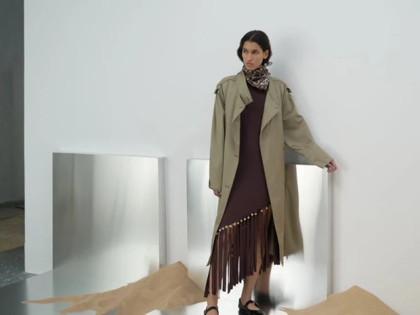 Fashion shot of a woman in a brown dress and tan trench coat standing in a studio with reflective decor and sand details on the floor.
