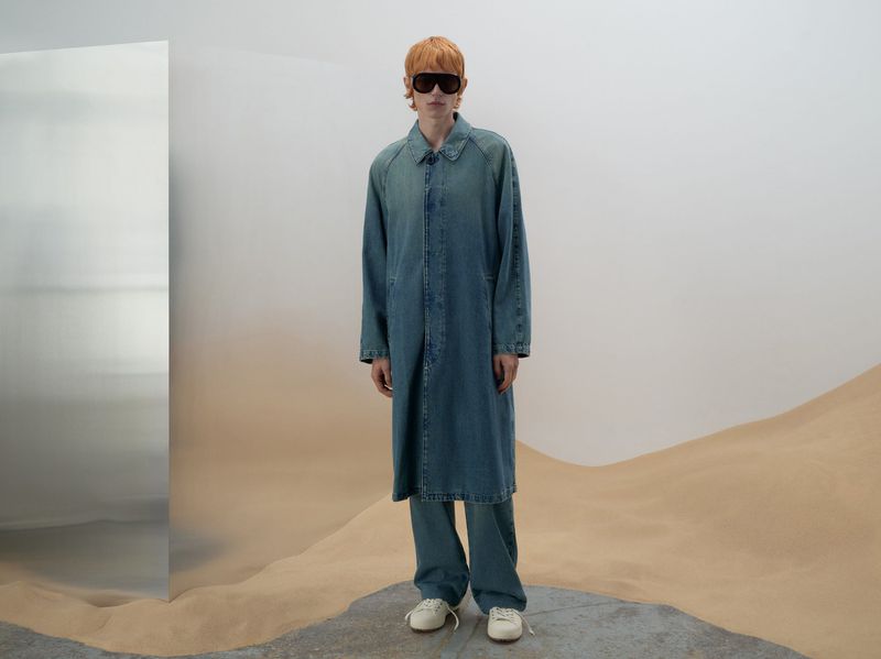 Person in a full-length denim coat and pants stands on a concrete surface with a tan desert-like backdrop and a silver reflective panel.