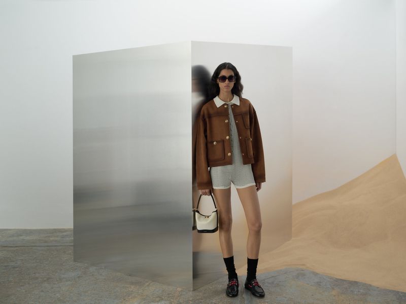 A woman posing in a brown jacket and matching shorts with a handbag stands indoors next to a reflective metal panel.