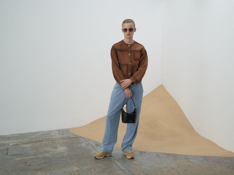 Adult poses in a brown jacket, light blue pants, and sunglasses, holding a black purse, standing indoors near a geometric design.